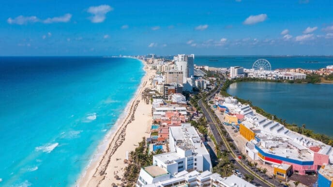 An expansive aerial view of Cancun, Mexico, showing a long stretch of white sand beach and turquoise ocean on the left, luxury hotels and resorts lining a narrow land strip, and a calm lagoon with a Ferris wheel on the right, all under a clear blue sky.