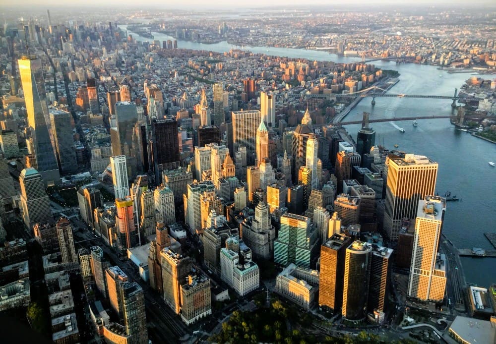 An expansive aerial view of the New York City skyline, with the sun's warm light reflecting off the numerous skyscrapers of Lower Manhattan, bordered by rivers, under a hazy sky.