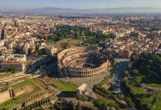 Aerial view of the Colosseum in Rome, Italy, surrounded by ancient ruins, city buildings, and distant mountains under a clear sky.