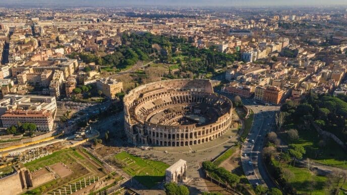 Aerial view of the Colosseum in Rome, Italy, surrounded by ancient ruins, city buildings, and distant mountains under a clear sky.