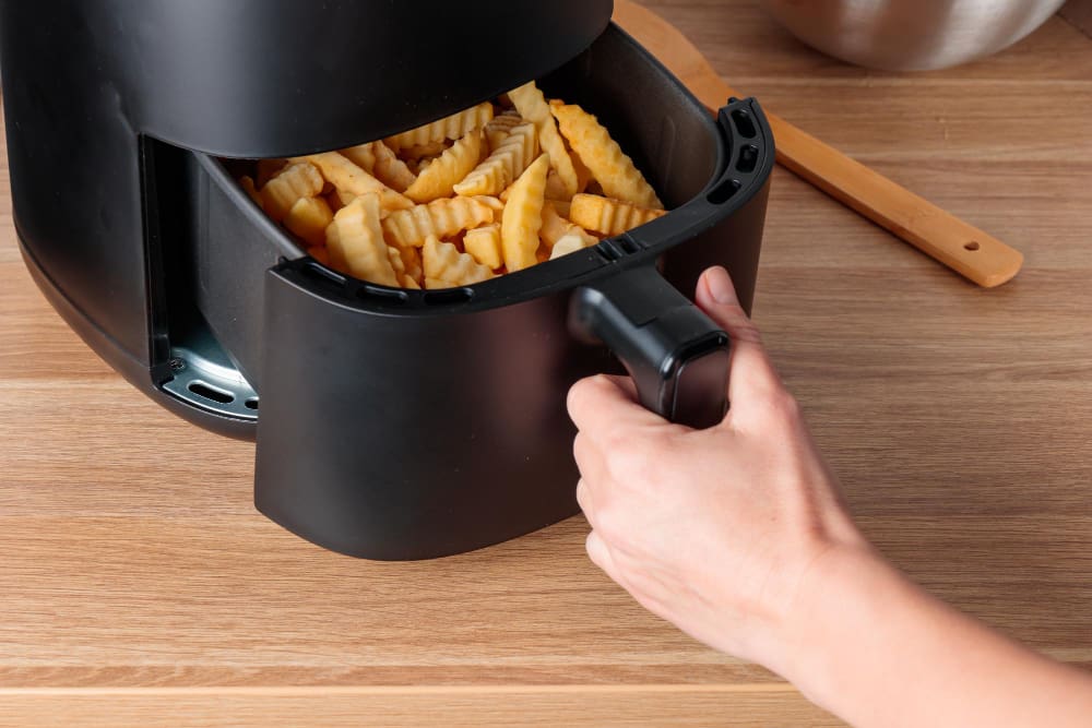 A hand pulling out the basket of an air fryer filled with golden, crispy crinkle-cut fries on a wooden countertop.