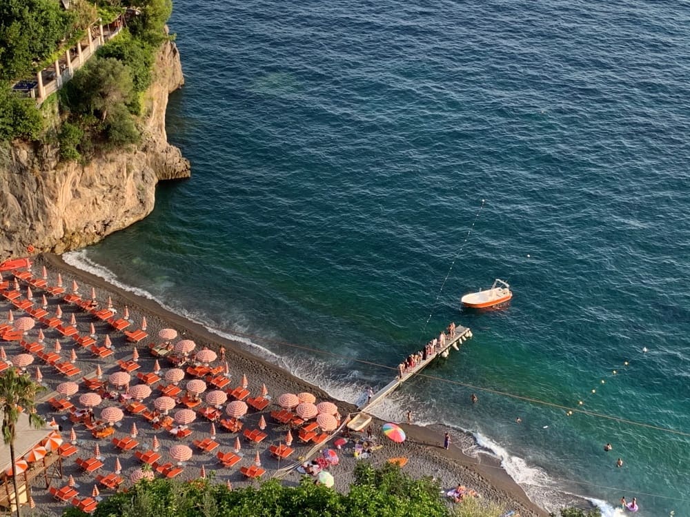 A high-angle view of a pebbly beach on the Amalfi Coast, Italy, covered in rows of orange-striped umbrellas and lounge chairs. A wooden pier extends into the clear turquoise sea, with steep cliffs rising on one side.