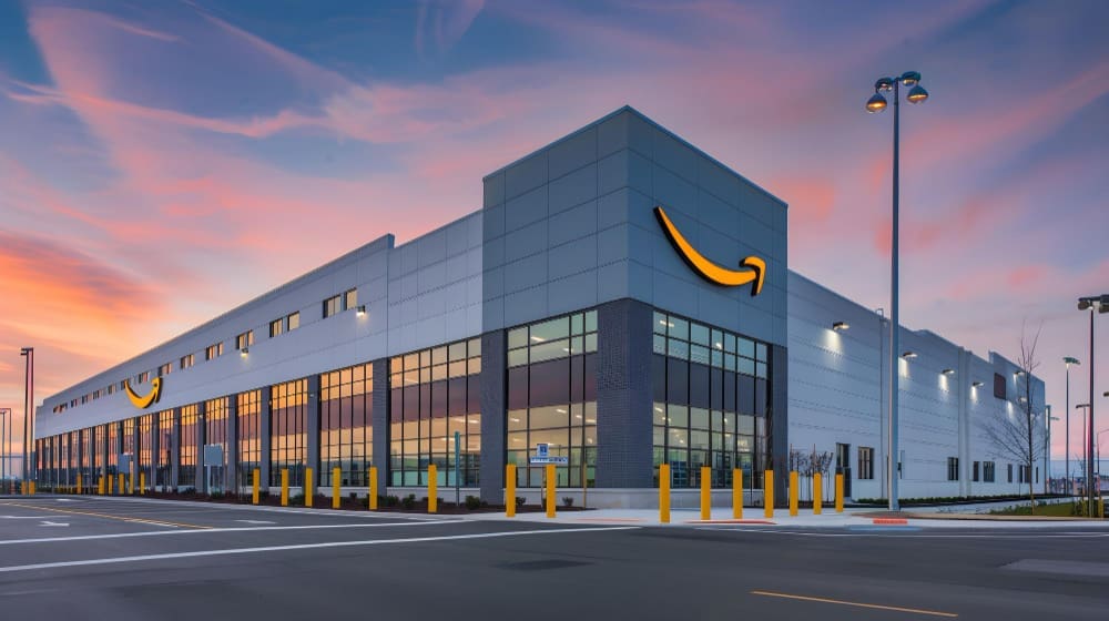 An Amazon fulfillment center building with its signature smile logo, under a colorful sunset sky.