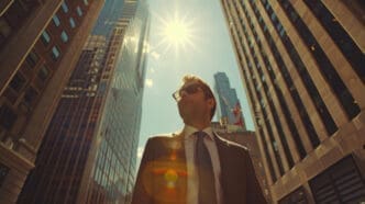 Low-angle shot of a man in a suit and sunglasses looking up confidently amidst towering city skyscrapers with a bright sun.