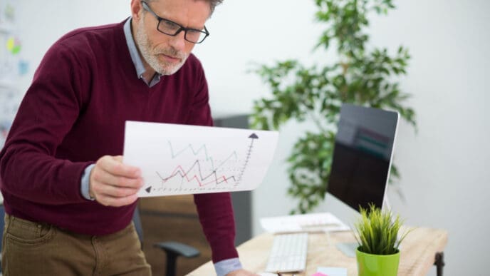 A man with grey hair and glasses, wearing a maroon sweater and khaki pants, stands at a desk, intently examining a paper with a line graph showing upward trends, with a computer monitor and plants in the background.