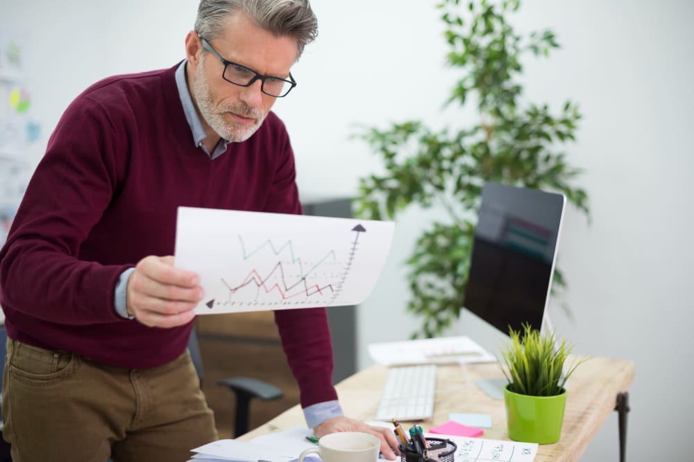 A man with grey hair and glasses, wearing a maroon sweater and khaki pants, stands at a desk, intently examining a paper with a line graph showing upward trends, with a computer monitor and plants in the background.