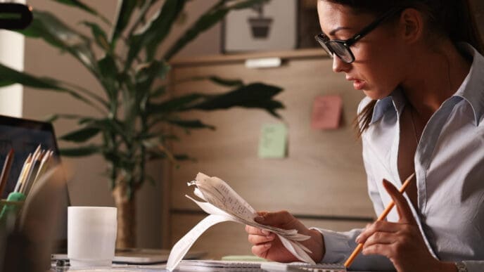 A woman wearing glasses is seen in profile, intently reviewing a stack of crumpled receipts in one hand while holding a pencil in the other, with a calculator and laptop on her desk.