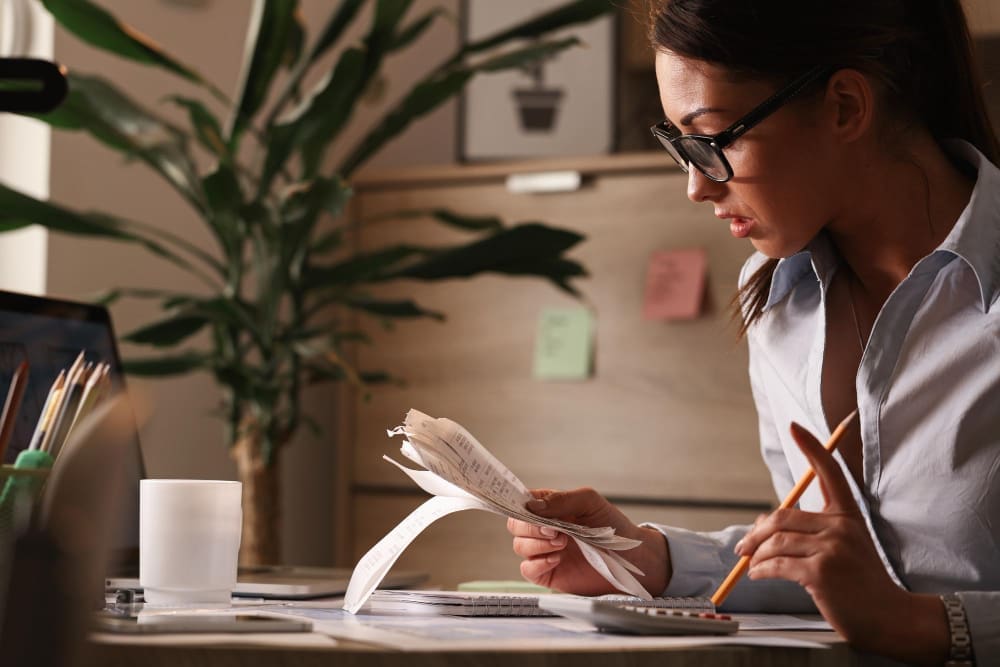 A woman wearing glasses is seen in profile, intently reviewing a stack of crumpled receipts in one hand while holding a pencil in the other, with a calculator and laptop on her desk.