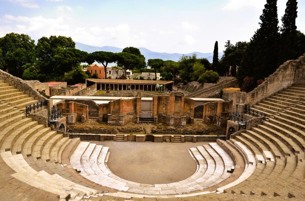 A high-angle view looking down into the ancient Roman Amphitheater of Pompeii, Italy, showing its stone seating tiers, central arena, and brick ruins, with modern buildings and mountains in the background under a cloudy sky.