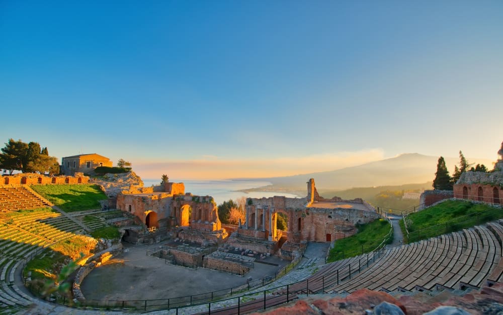 A wide-angle view of the ancient Greek Theatre of Taormina in Sicily, Italy, showing its stone seating tiers and ruins, with the sea and distant Mount Etna visible under a golden sunrise or sunset sky.