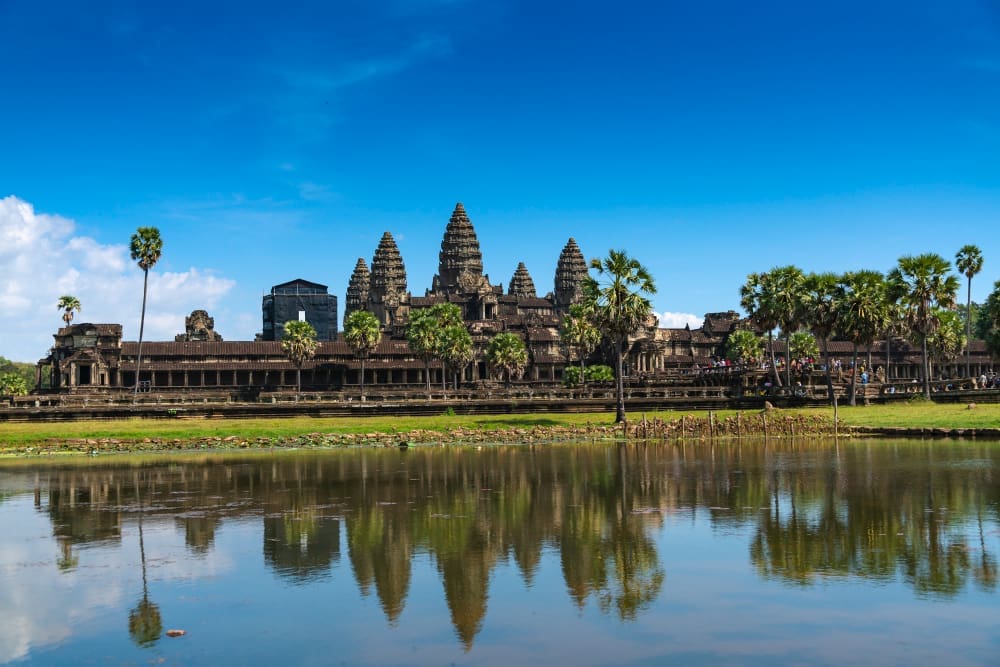 The majestic Angkor Wat temple complex, with its distinctive towers and intricate stone architecture, is reflected in the calm water of a foreground pond, surrounded by palm trees under a bright blue sky.