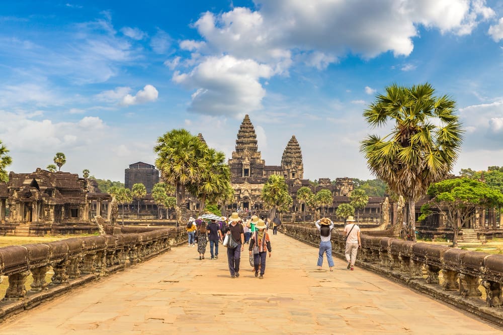 Tourists walking on a causeway towards the grand Angkor Wat temple in Siem Reap, Cambodia, surrounded by palm trees under a blue sky.