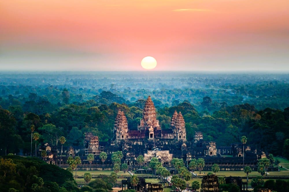 Aerial view of Angkor Wat temple complex, surrounded by dense forest, under a glowing sunrise sky.