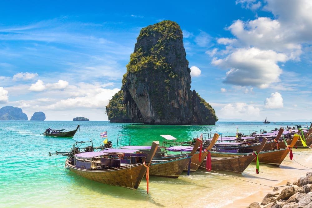 Longtail boats lined up on the sandy shore of Ao Phra Nang Beach in Krabi, Thailand, with towering limestone karsts and turquoise water under a blue sky.