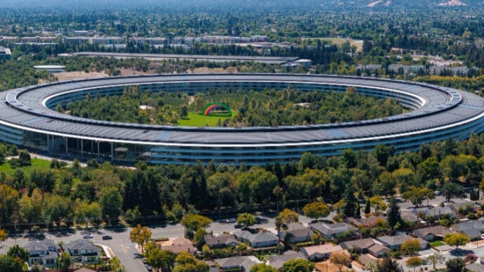An aerial view of Apple Park in Cupertino, California, showing the massive circular "spaceship" campus surrounded by abundant trees and suburban housing, with distant hills under a clear blue sky.