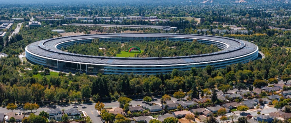 An aerial view of Apple Park in Cupertino, California, showing the massive circular "spaceship" campus surrounded by abundant trees and suburban housing, with distant hills under a clear blue sky.