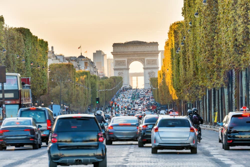 A wide, tree-lined Avenue des Champs-Élysées in Paris is seen from behind a busy flow of cars, leading to the majestic Arc de Triomphe in the distance, bathed in golden light.