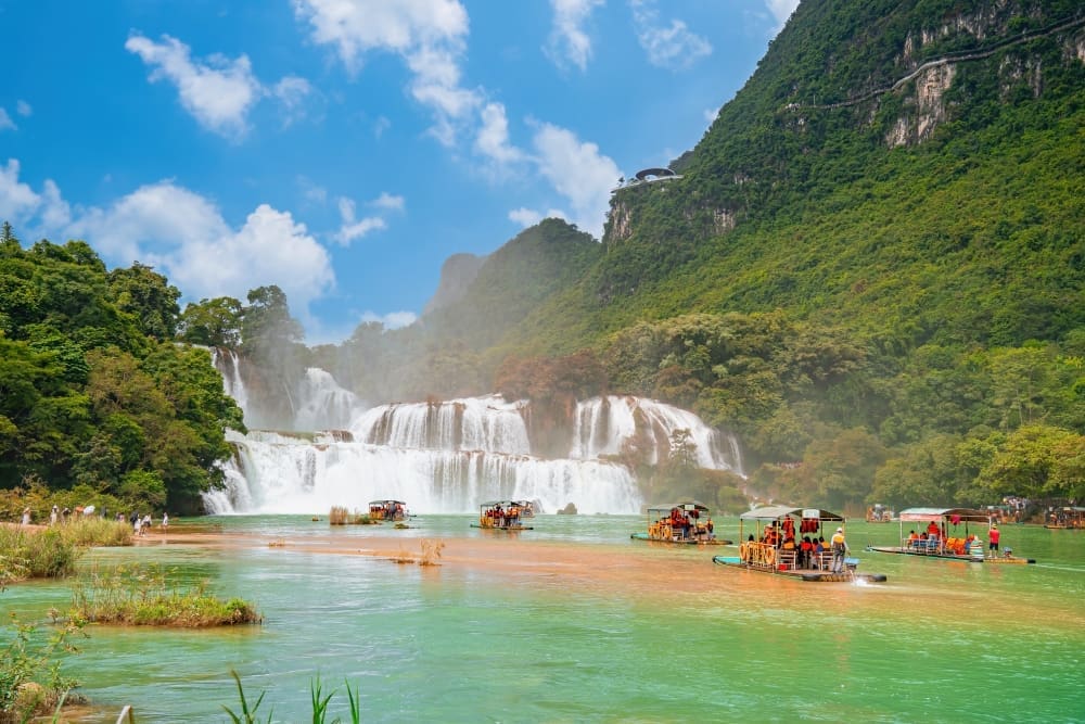 The majestic Ban Gioc Waterfall in Cao Bang, Vietnam, cascading into a turquoise river with bamboo rafts carrying tourists, surrounded by lush mountains.