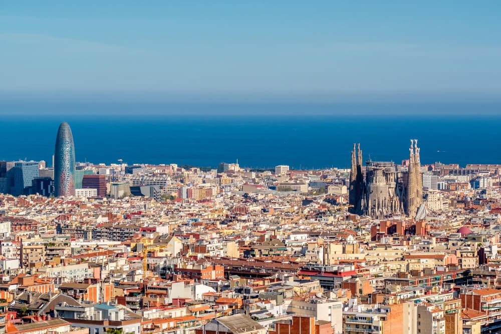 Panoramic view of Barcelona's cityscape with Sagrada Familia, Agbar Tower, and the Mediterranean Sea in the background.