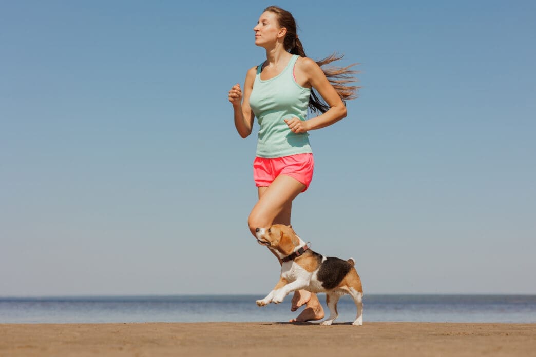 Young woman in activewear running on a beach with a beagle.