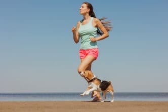 Young woman in activewear running on a beach with a beagle.