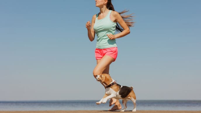 Young woman in activewear running on a beach with a beagle.
