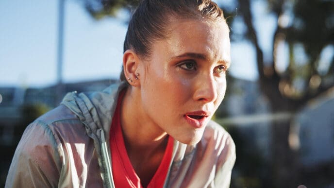A close-up of a young woman's face, covered in sweat, with her mouth slightly open and eyes wide, looking determinedly after an intense run outdoors.
