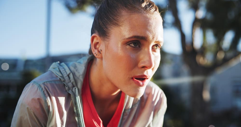 A close-up of a young woman's face, covered in sweat, with her mouth slightly open and eyes wide, looking determinedly after an intense run outdoors.