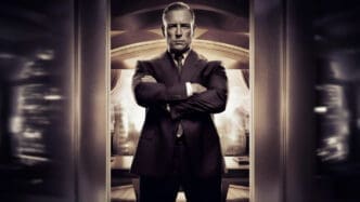 A serious man in a dark suit with slicked-back hair stands with arms crossed, framed by a doorway, with a blurred, stylized city backdrop and a large star symbol above, evoking power and dynasty.
