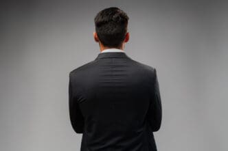 The back of a man's head and shoulders, wearing a dark suit, facing away from the viewer against a plain grey background, symbolizing leadership, mystery, or a forward-looking stance.