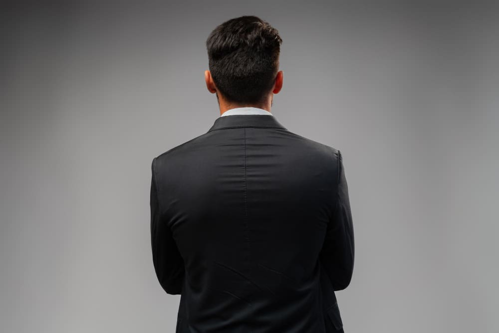The back of a man's head and shoulders, wearing a dark suit, facing away from the viewer against a plain grey background, symbolizing leadership, mystery, or a forward-looking stance.