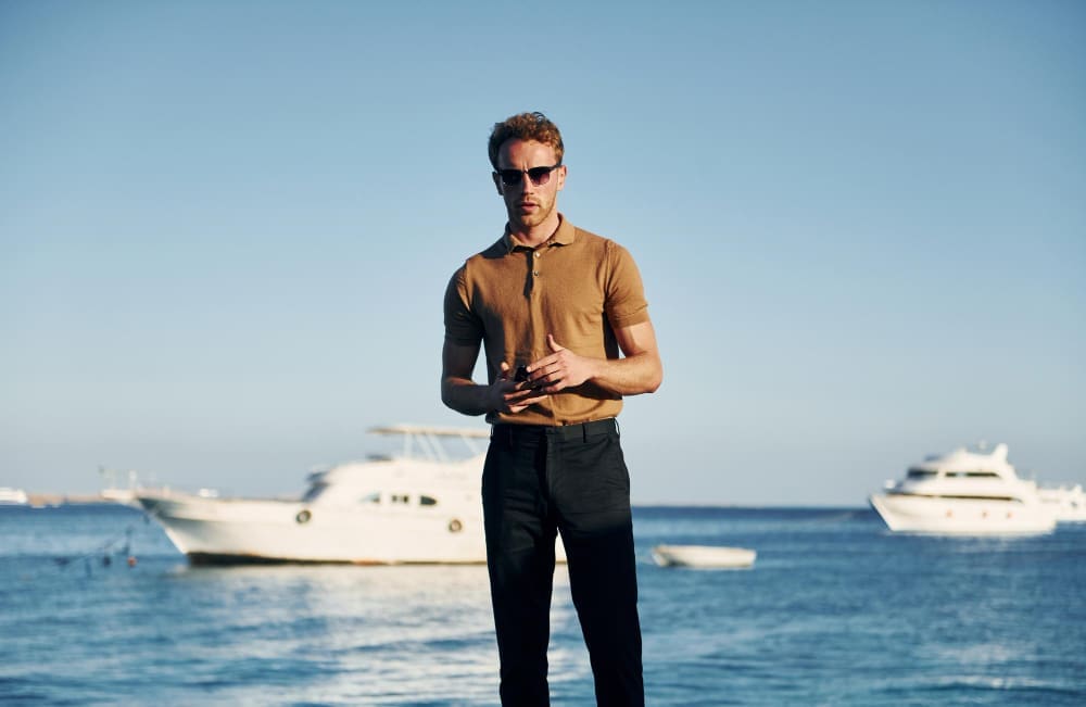 A man in a brown polo shirt and black pants stands on a dock, wearing sunglasses and clasping his hands, with luxury yachts on the calm blue water and a clear sky in the background.