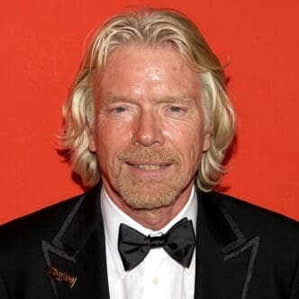 A headshot of Richard Branson, with long blonde hair and a beard, smiling faintly while wearing a black tuxedo and bowtie with a "TIME 100" pin, against a vibrant red-orange background.
