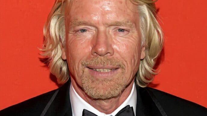 A headshot of Richard Branson, with long blonde hair and a beard, smiling faintly while wearing a black tuxedo and bowtie with a "TIME 100" pin, against a vibrant red-orange background.