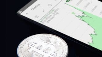 A silver physical Bitcoin coin rests on a dark surface, with a smartphone in the background displaying a green-lined cryptocurrency chart showing an upward trend.