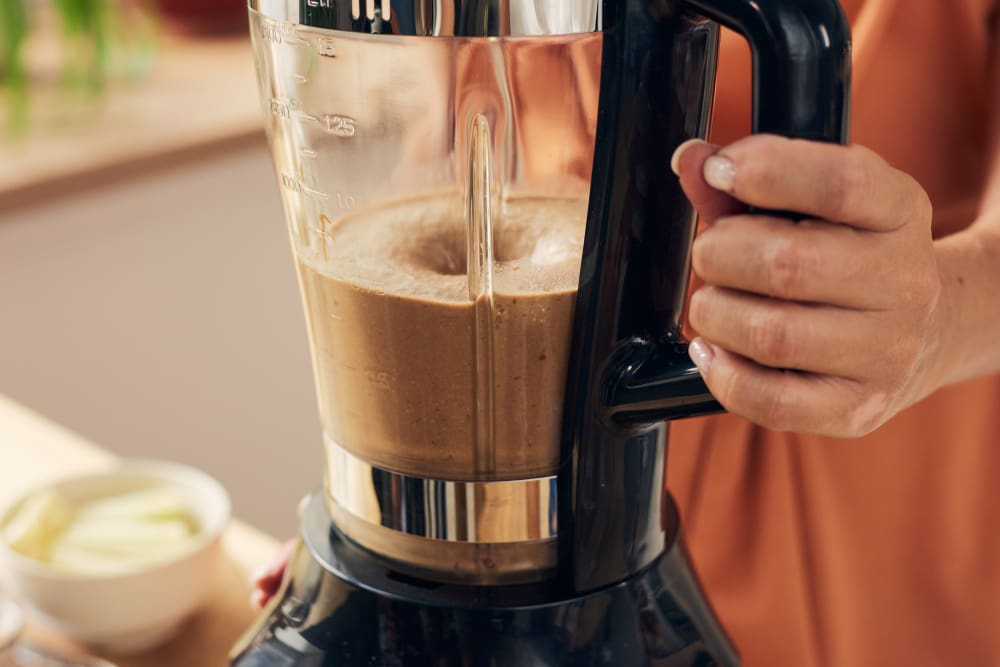 A close-up of a blender mixing a brown liquid, held by a person's hand.