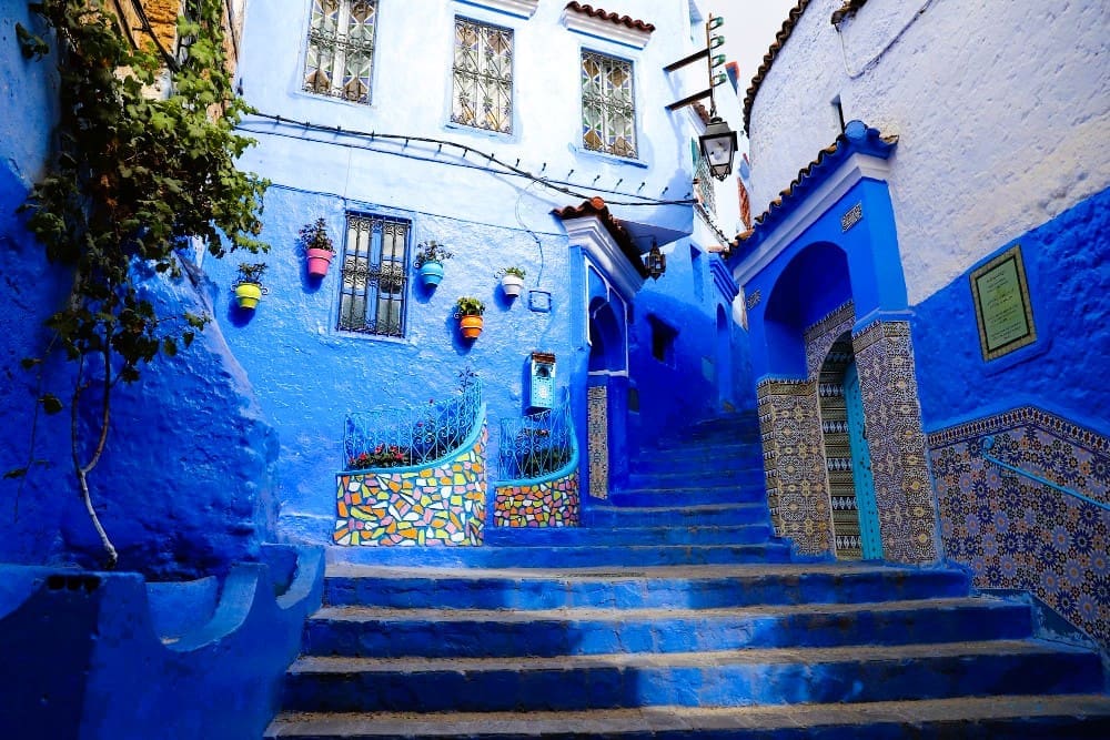 A narrow, vibrant blue street in Chefchaouen, Morocco, with stairs, potted plants, and ornate doorways.