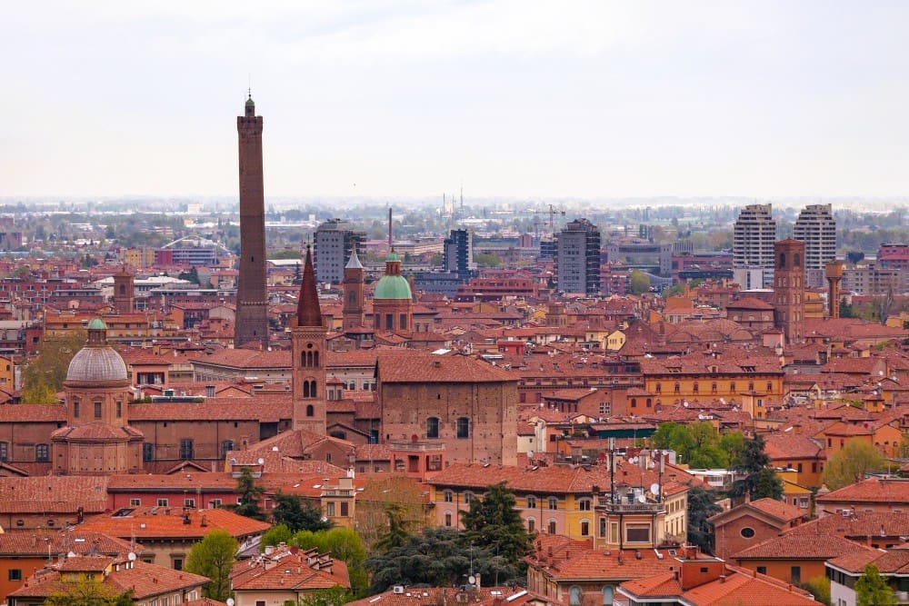 An elevated panoramic view of Bologna, Italy, showcasing its dense cityscape of terracotta rooftops, prominent medieval towers (including a leaning one), and numerous church domes under a hazy sky.