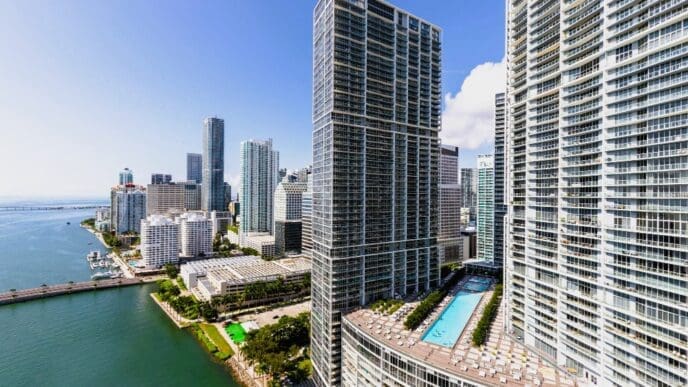 A high-angle view of the dense, modern skyline of Brickell, Miami, with tall skyscrapers and a rooftop swimming pool, overlooking a bay with a bridge, under a bright blue sky with scattered clouds.