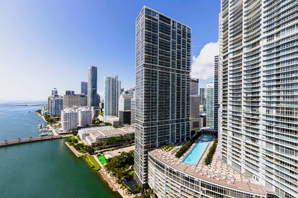 A high-angle view of the dense, modern skyline of Brickell, Miami, with tall skyscrapers and a rooftop swimming pool, overlooking a bay with a bridge, under a bright blue sky with scattered clouds.