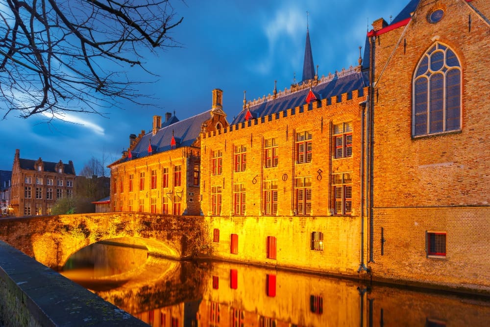 The illuminated historic building of Brugse Vrije and a stone bridge reflecting in the Green Canal in Bruges at night.