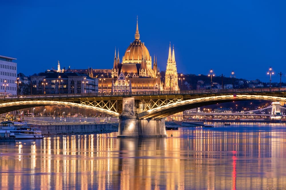 The illuminated Hungarian Parliament Building and Széchenyi Chain Bridge reflecting in the Danube River at dusk in Budapest.