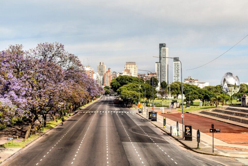 A wide, tree-lined avenue with purple jacaranda blossoms in Buenos Aires, Argentina, leading towards a modern city skyline with skyscrapers and the Floralis Genérica sculpture.