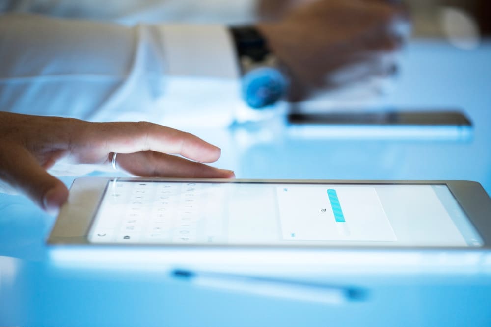 A hand interacts with a tablet showing a loading bar, placed on a glowing blue surface, with blurred hands and a smartphone in the background, symbolizing data processing and readiness for AI.