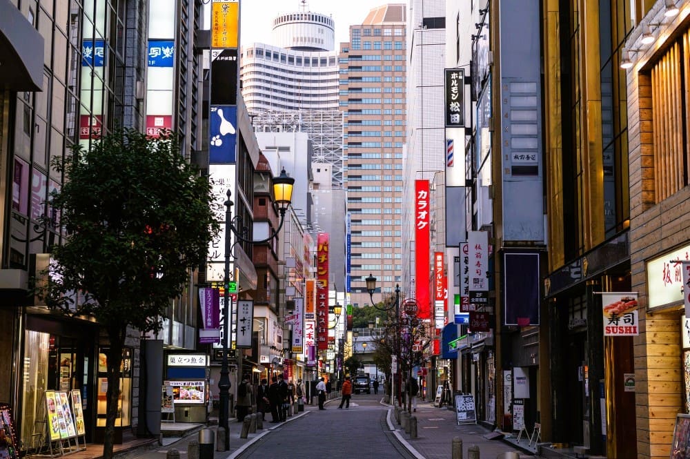 A bustling street scene in Osaka, Japan, lined with tall buildings adorned with numerous vertical and horizontal signs in Japanese characters, with pedestrians in the distance and modern skyscrapers in the background.