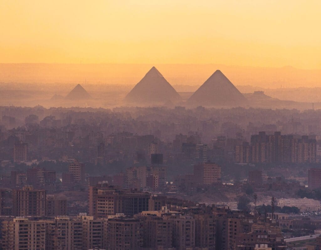 An aerial view of a sprawling city with the Giza Pyramids visible in the hazy distance against a sunset sky.