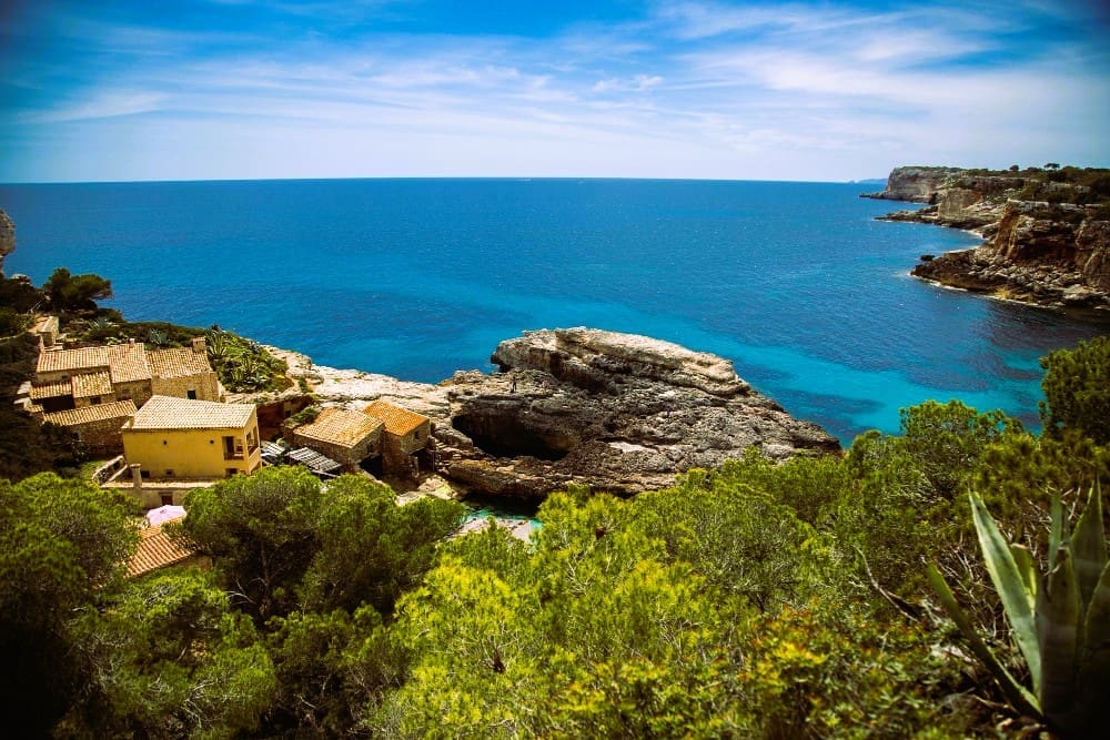 A high-angle panoramic view of a beautiful turquoise bay in Mallorca, Spain, with traditional stone houses nestled on the rocky coastline, surrounded by green trees, and distant cliffs under a clear blue sky.