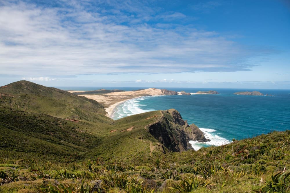 A panoramic coastal view of rugged green hills sloping down to a long sandy beach where turquoise waves meet the shore. The deep blue ocean extends to the horizon, dotted with small islands, under a partly cloudy blue sky in New Zealand.