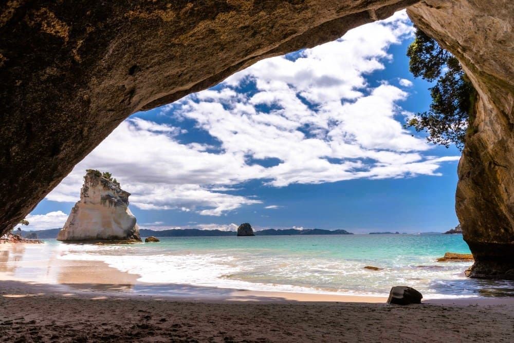 A stunning view through a large rock archway opening onto a sandy beach and turquoise ocean, with a distinctive sea stack and distant islands under a bright blue sky with white clouds at Cathedral Cove.