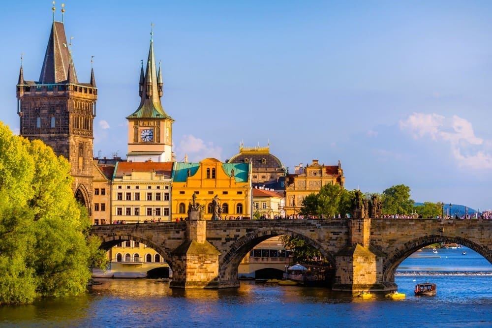 The Charles Bridge in Prague, adorned with statues and towers, spanning the Vltava River, with historic buildings under a blue sky.
